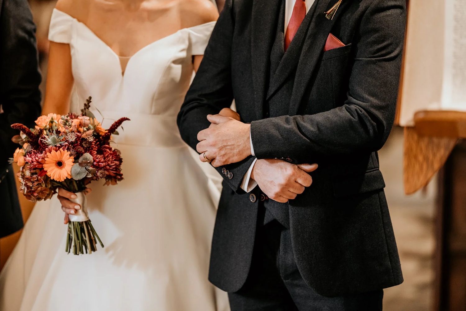 Closeup of wedding couple with white dress and black suit holding a vibrant autumn-colored bouquet in church.