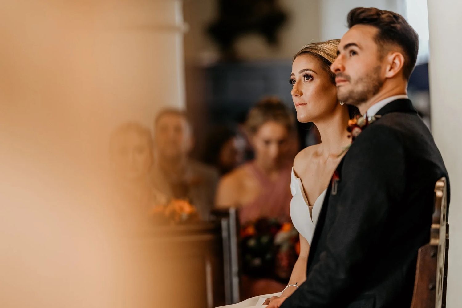 A couple in elegant wedding attire stands together in a warm-lit ceremony space.
