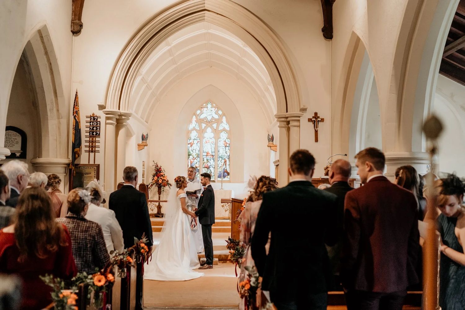 Wedding guests gather inside a church during a ceremony while an attendant lights candles along the aisle.