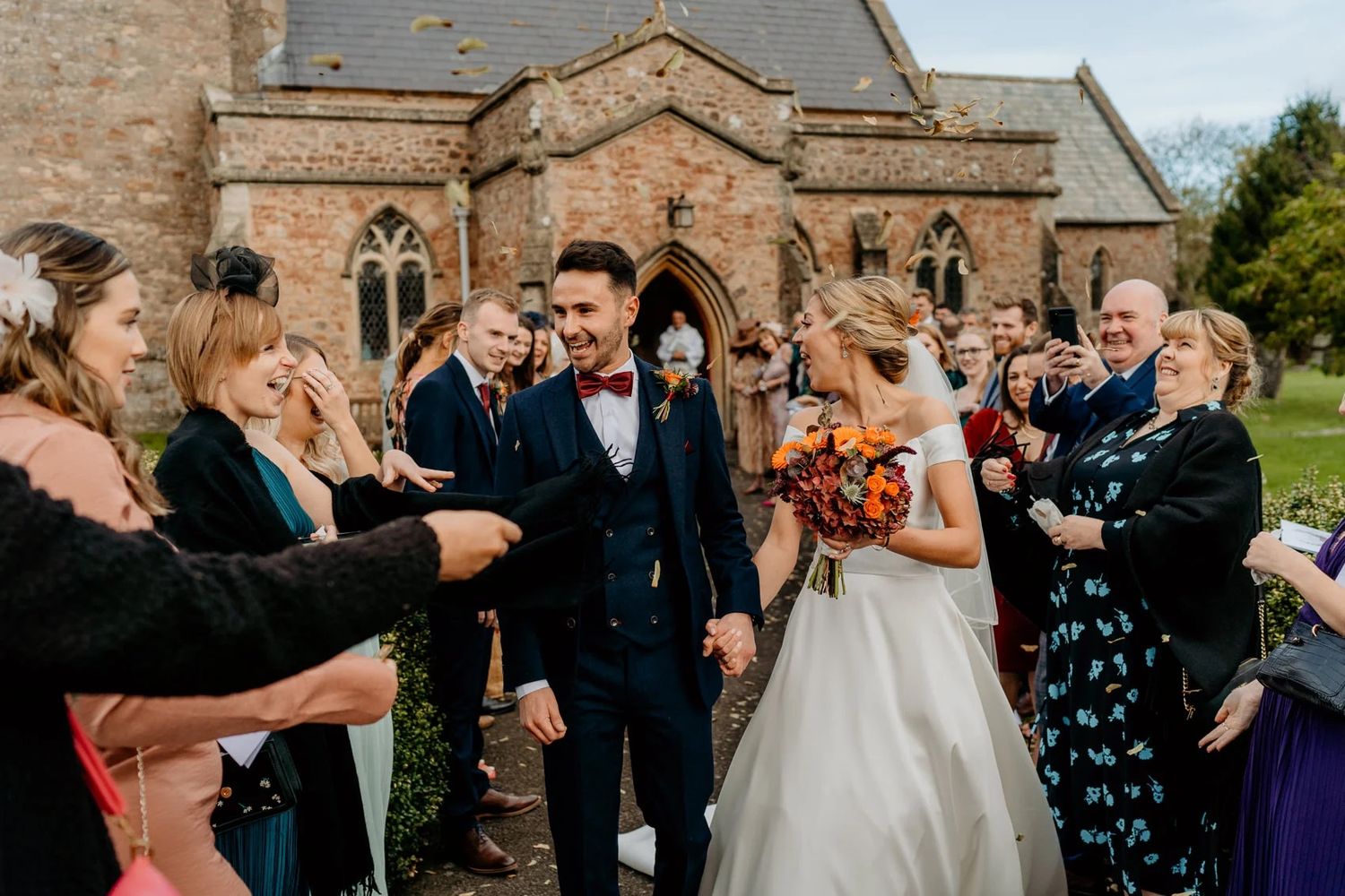 Guests celebrate and throw confetti outside historic stone church during wedding celebration.