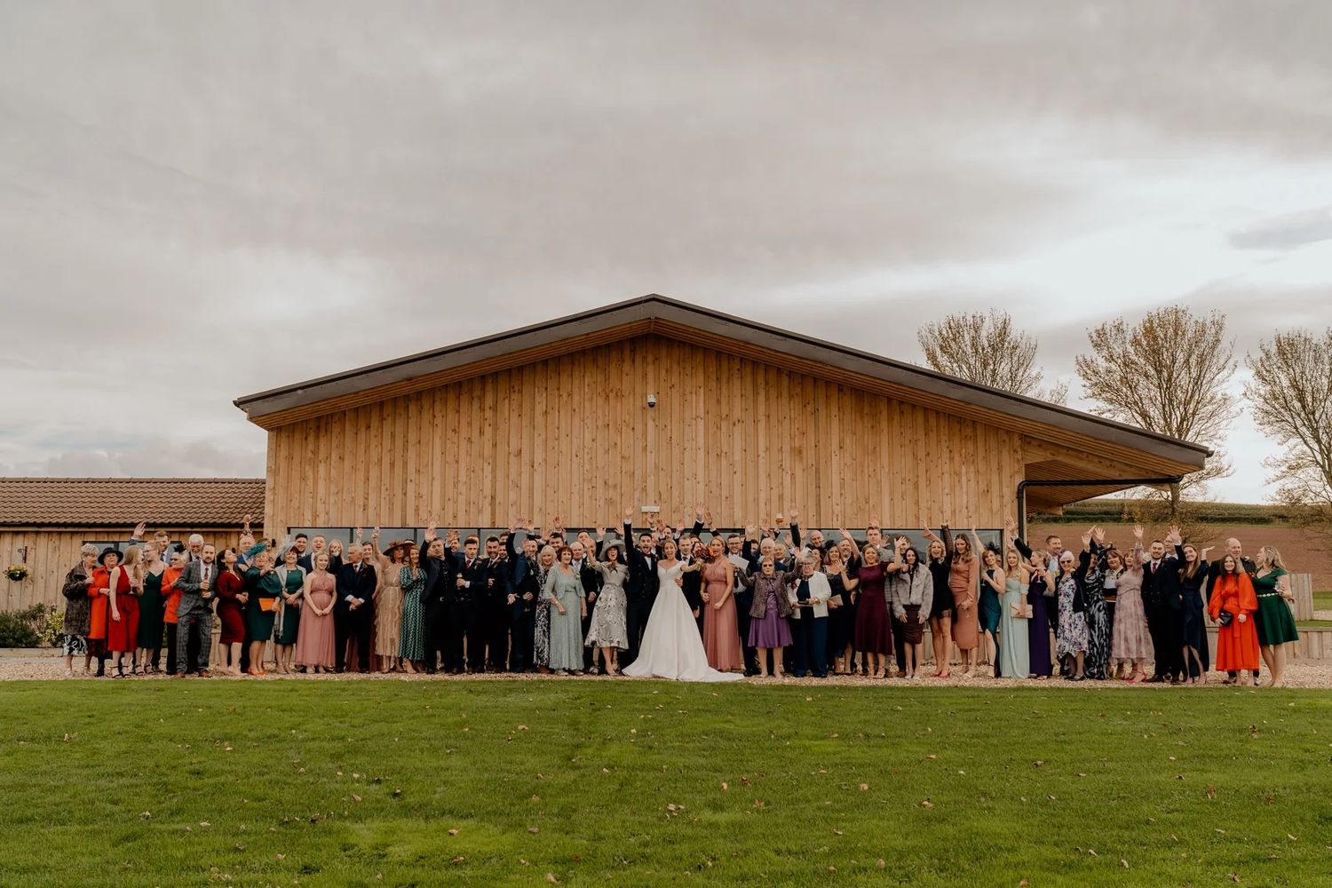 Large group wedding photo of guests and couple in front of modern wooden barn venue.