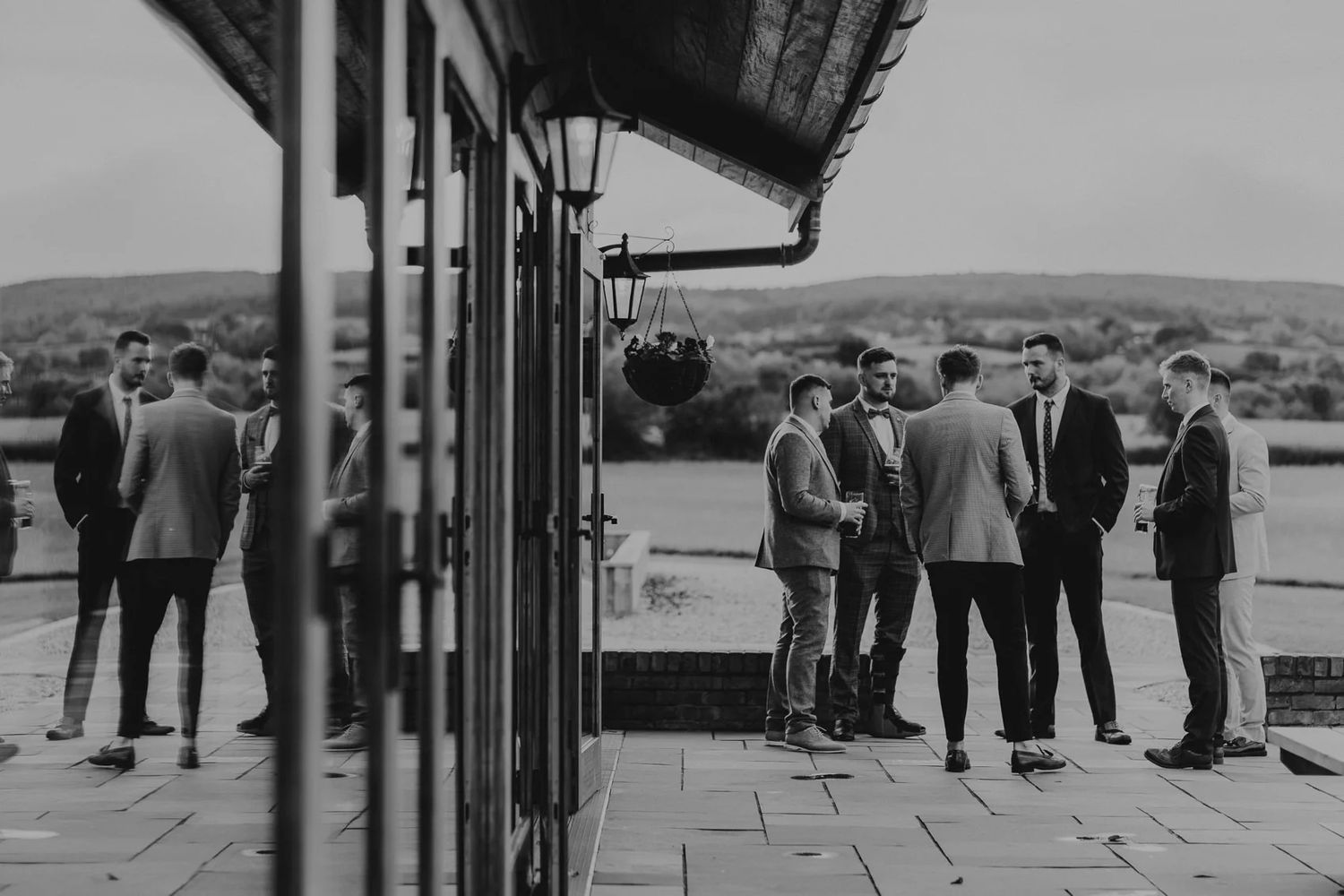 Black and white photo of wedding guests gathered on outdoor terrace with countryside view.