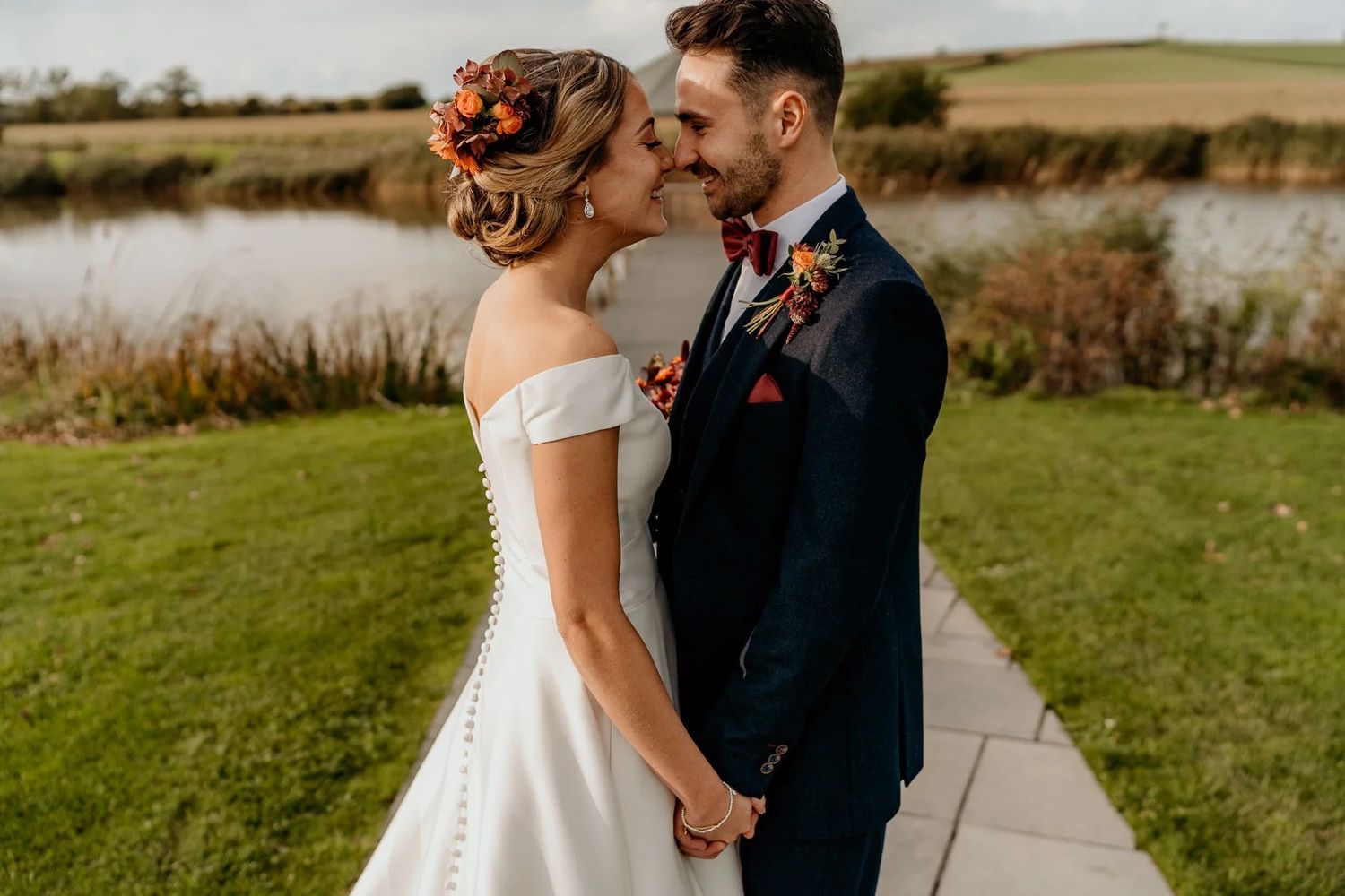 Romantic wedding couple standing in golden field by lake during sunset.