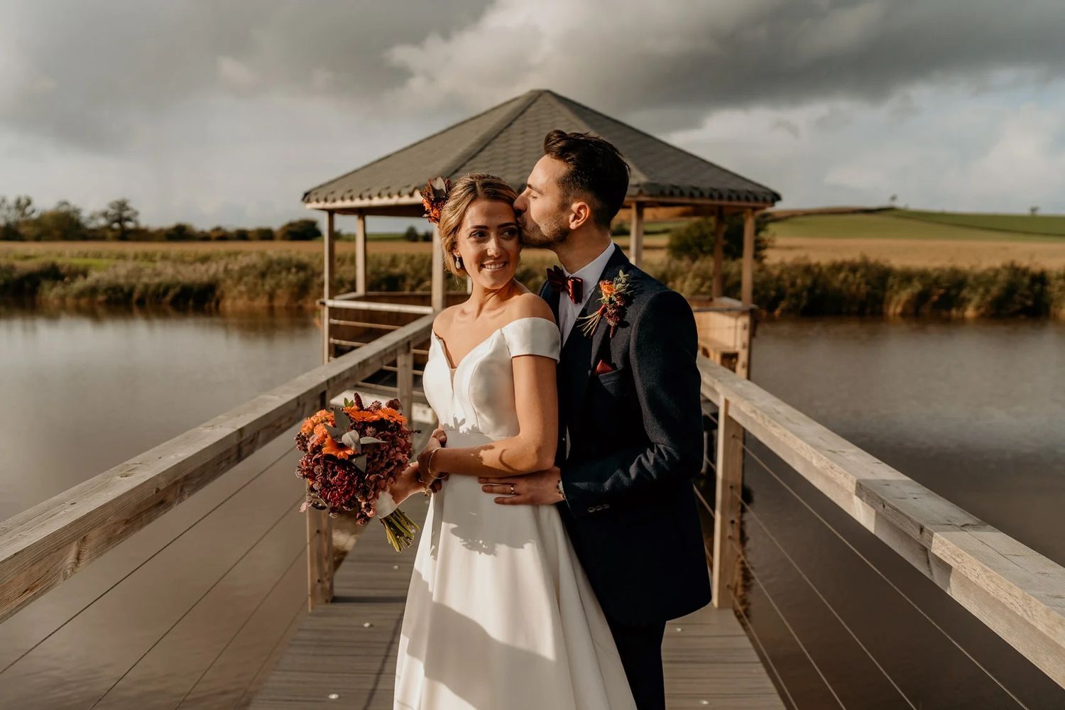 Wedding couple shares intimate moment on wooden dock overlooking water during sunset.