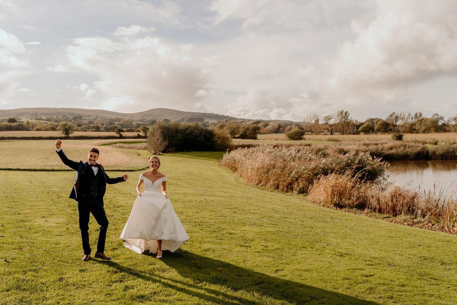 Wedding couple walks together through scenic countryside with golden sunlight across green fields.