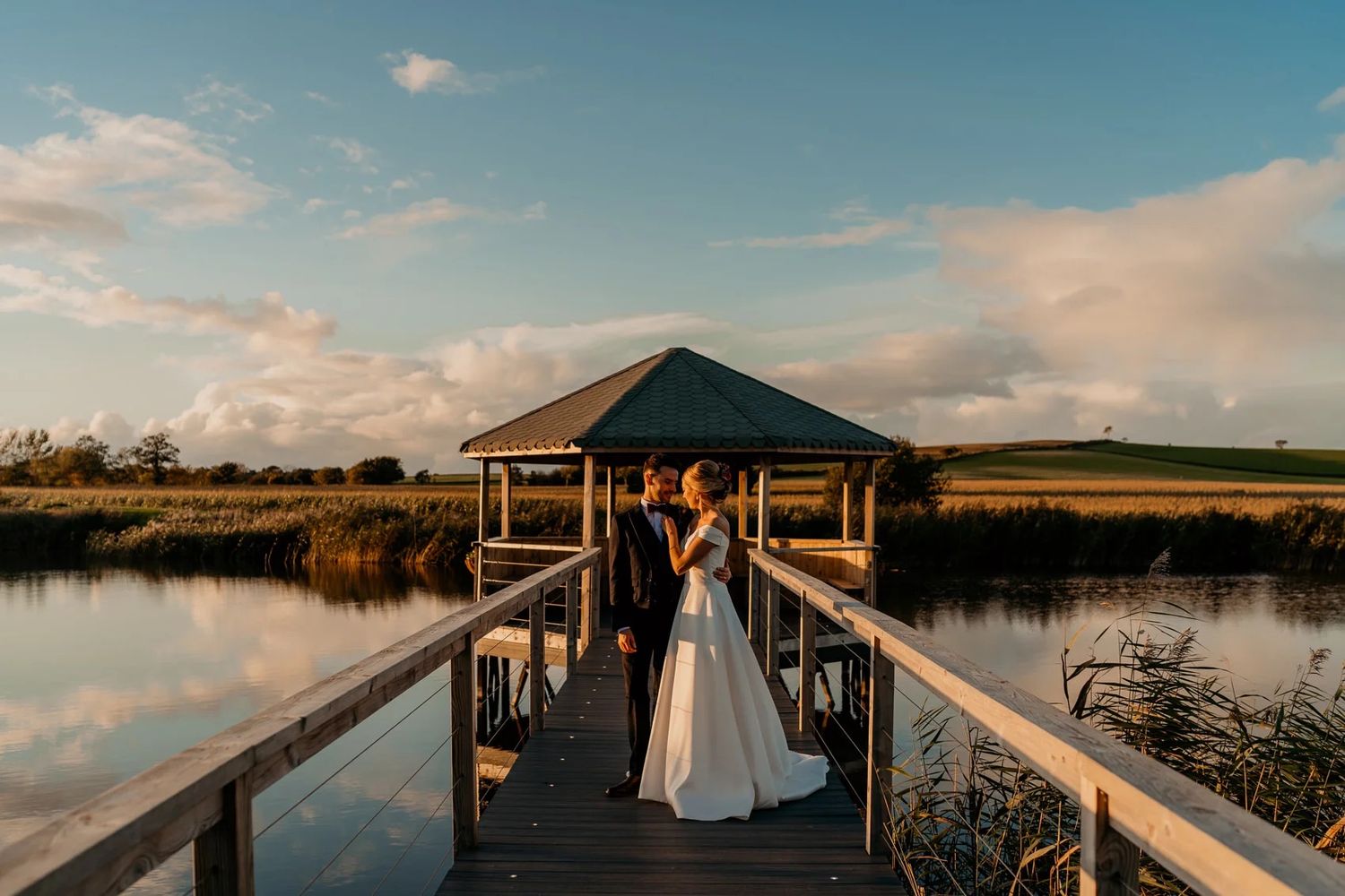 Wedding couple on wooden dock overlooking lake during golden hour.