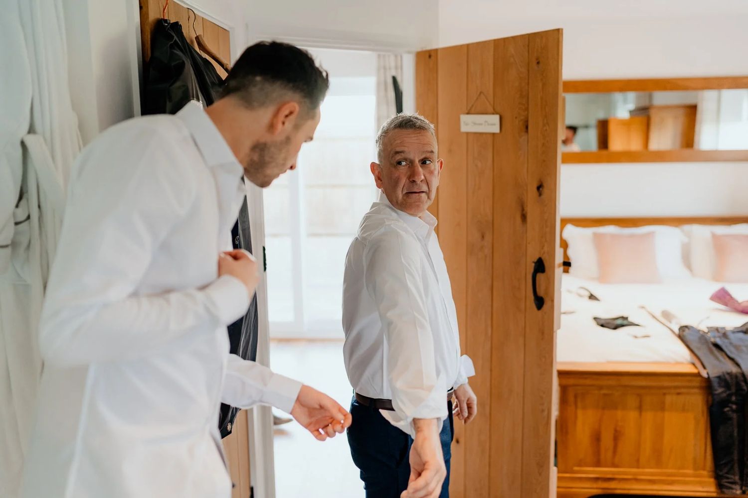 Two men in white dress shirts stand in a bright room with wooden furniture and storage.