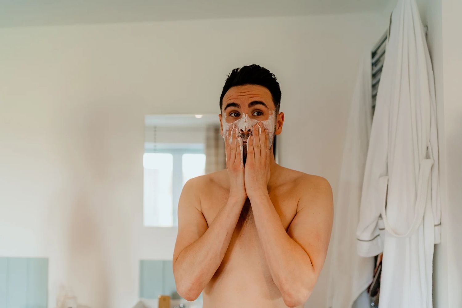 Person with black hair stands shirtless in a brightly lit bathroom covering their mouth with both hands.