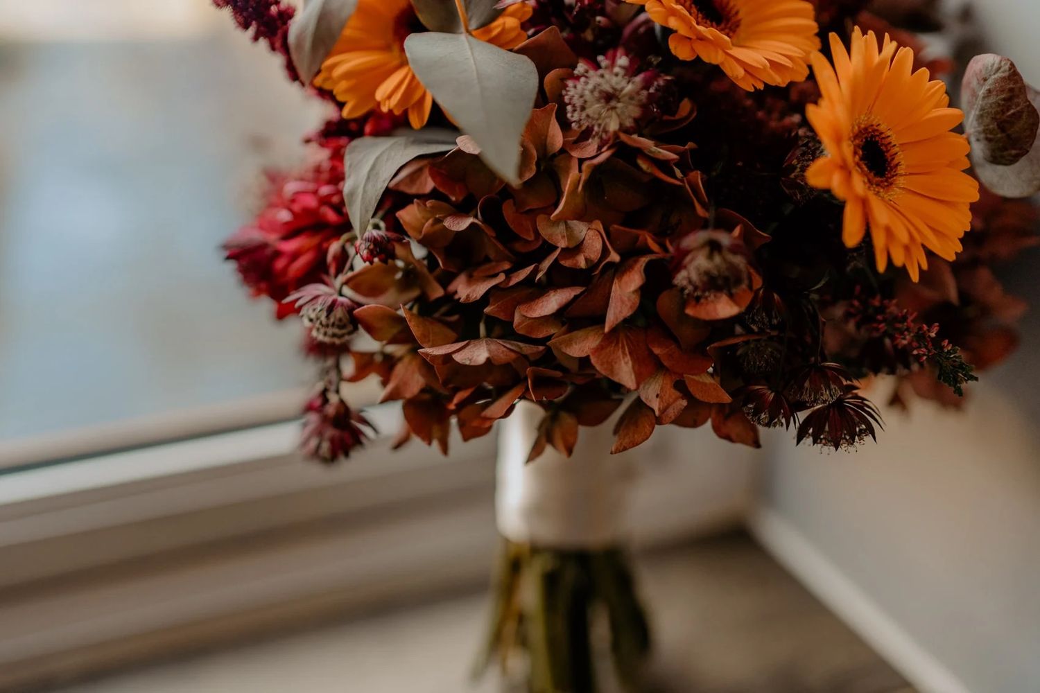 Orange and pink flower arrangements in vases line a window sill with sunlight streaming through.