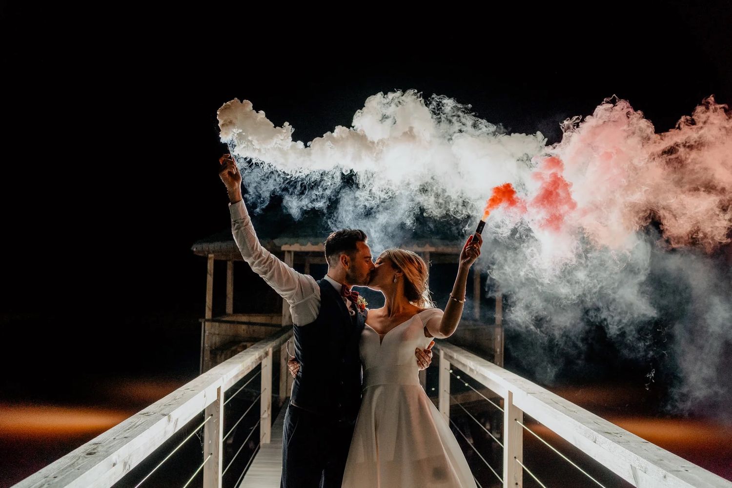 Wedding couple on a pier at night holding colorful smoke bombs creating dramatic red and white smoke effects against dark sky.