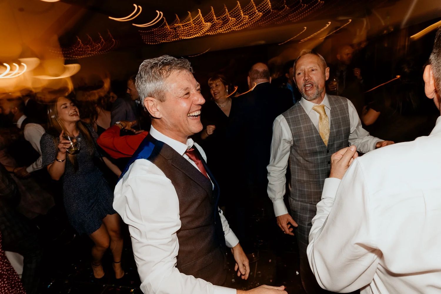 Wedding guests dance under warm lighting at a lively evening reception celebration.