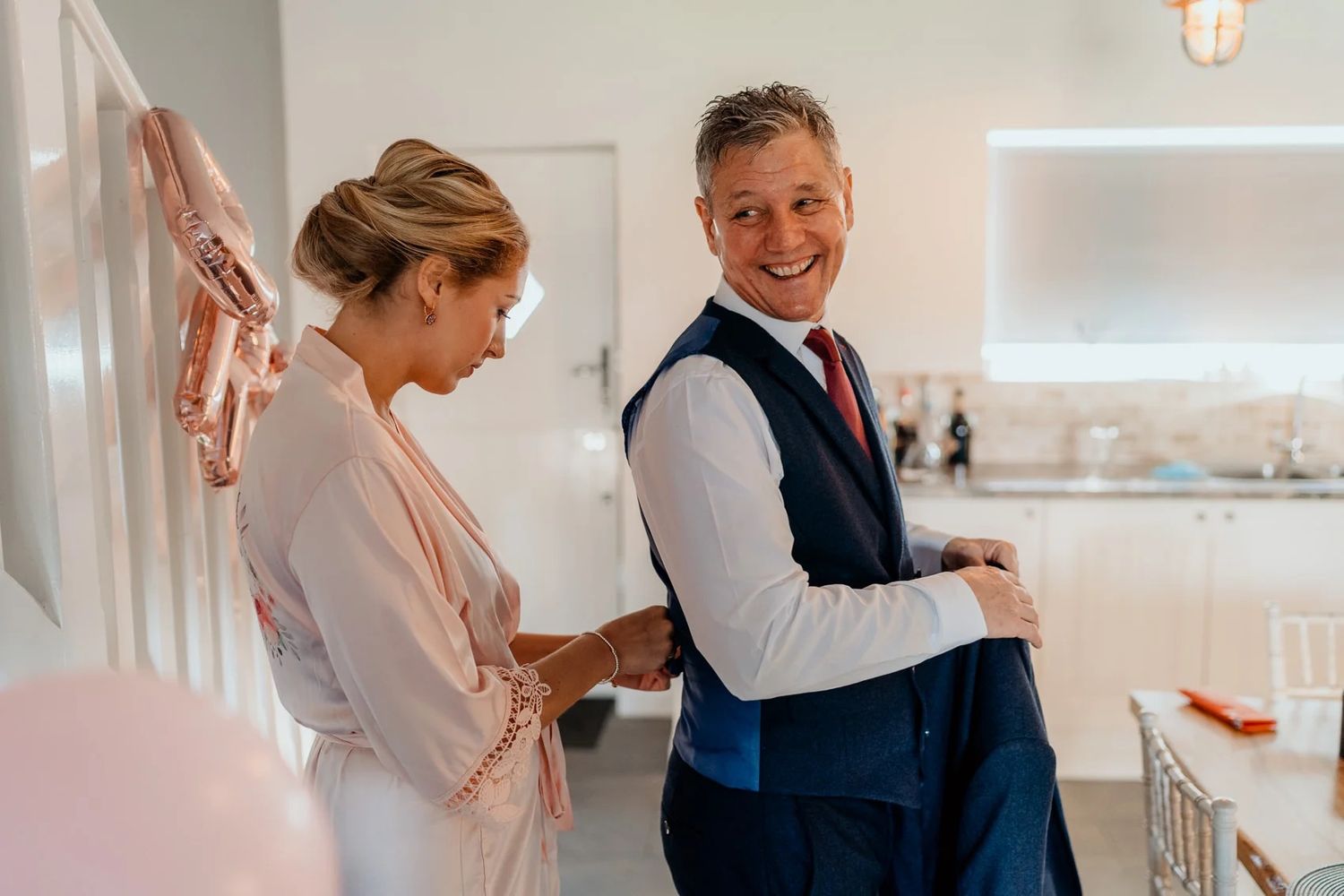 Wedding party members in dusty pink dresses help with final preparations in a bright sunlit room.