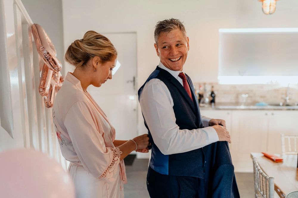 Wedding party members in dusty pink dresses help with final preparations in a bright sunlit room.