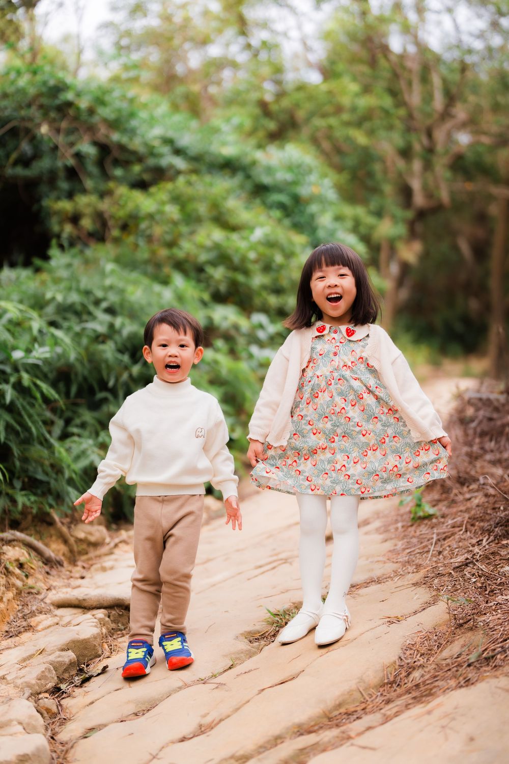 Two children laughing and playing together on a nature trail surrounded by lush green foliage on a sunny day.