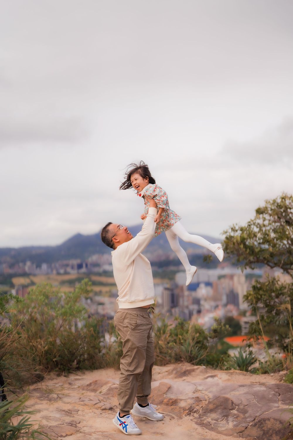 A father playfully holds his daughter on the top of Junjianyan Hiking Trail overlooking scenic Shilin District Taipei below