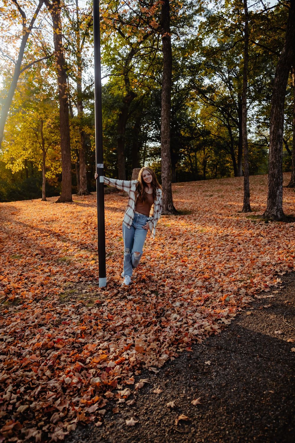Someone poses casually on a paved path surrounded by fallen autumn leaves and sunlit trees in a park setting.