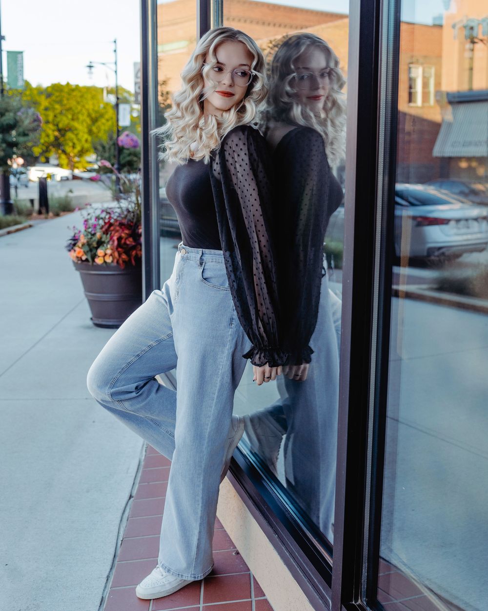Two women posing together in casual outfits with jeans and sweaters on a city sidewalk by a storefront window.