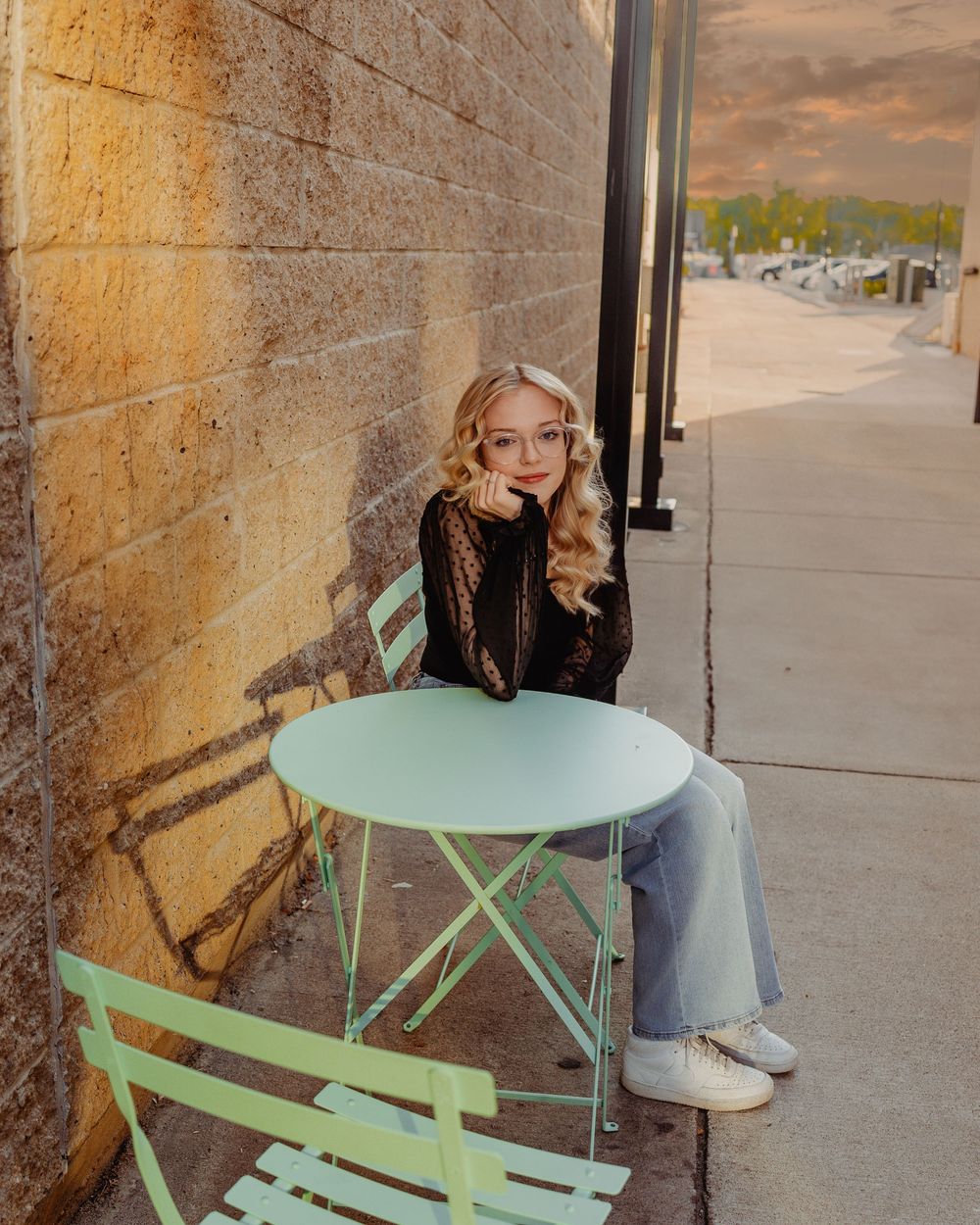 A person sits on a mint green bistro chair against a stone wall during golden hour lighting on a sidewalk.