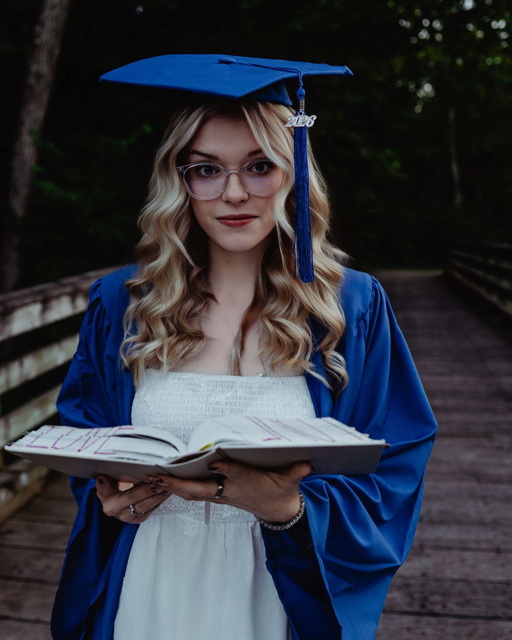 Graduate student in blue cap and gown poses with a book on a wooden bridge in an outdoor setting.