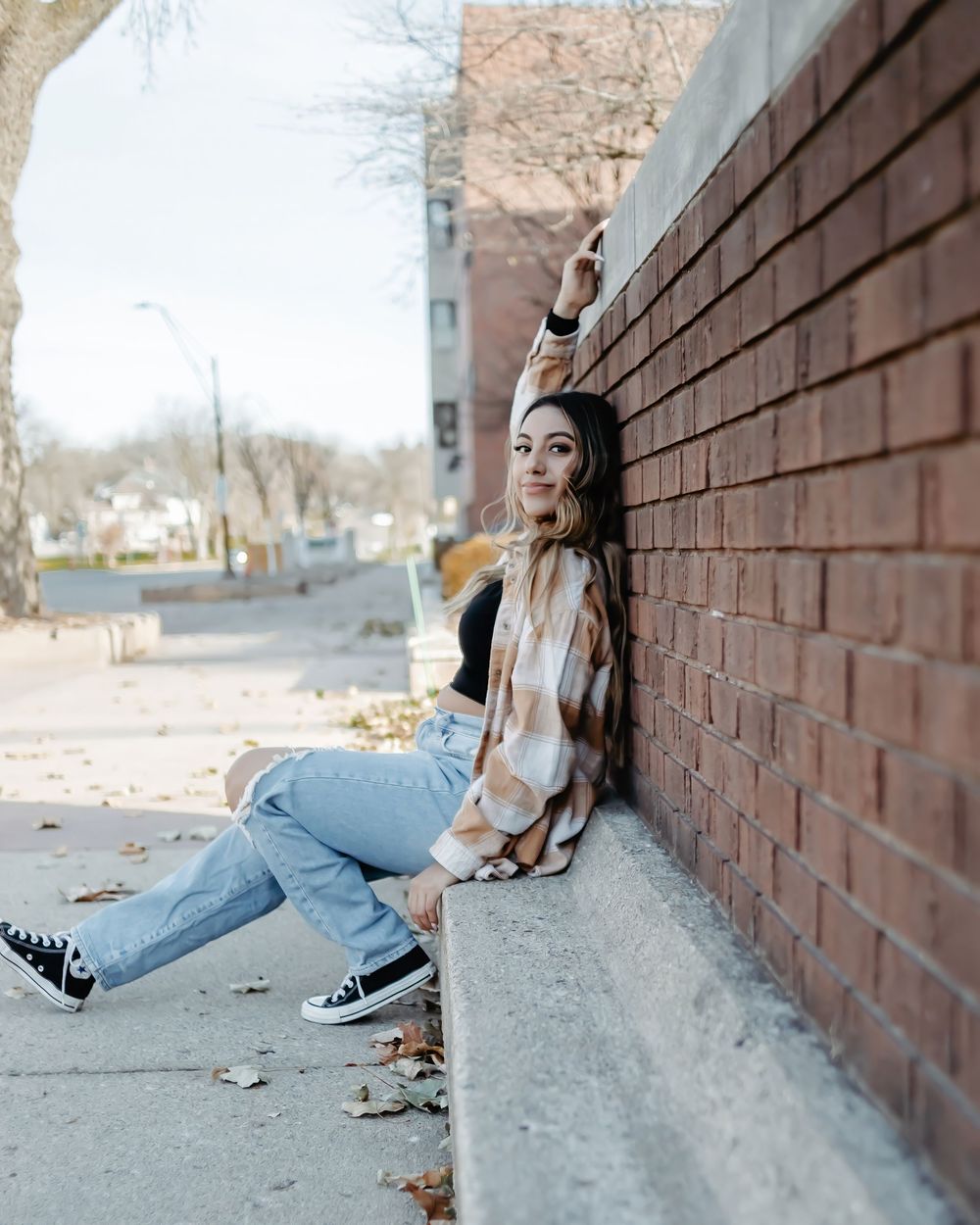 A person in casual attire and jeans rests against a brick wall in an outdoor urban setting on a fall day.