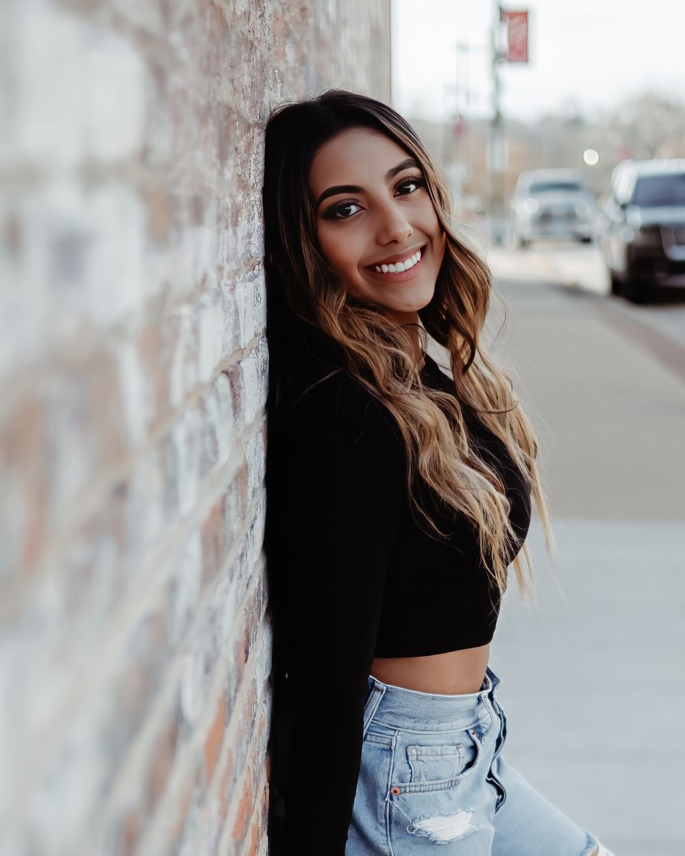 A fashionable woman in a black crop top and ripped jeans posing casually against a textured concrete wall.