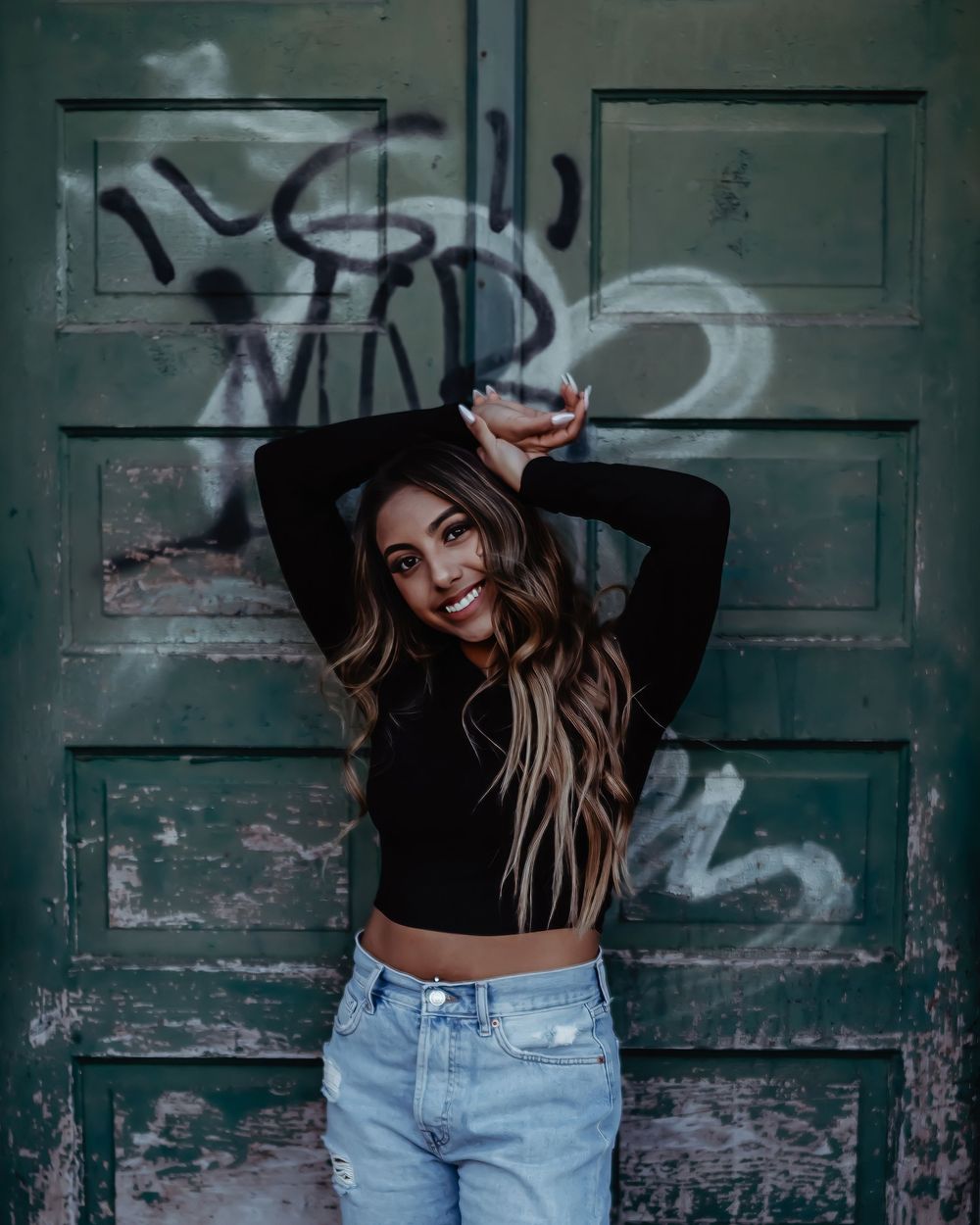 Portrait against a rustic green door with graffiti showing someone in a black crop top and light denim jeans posing playfully.