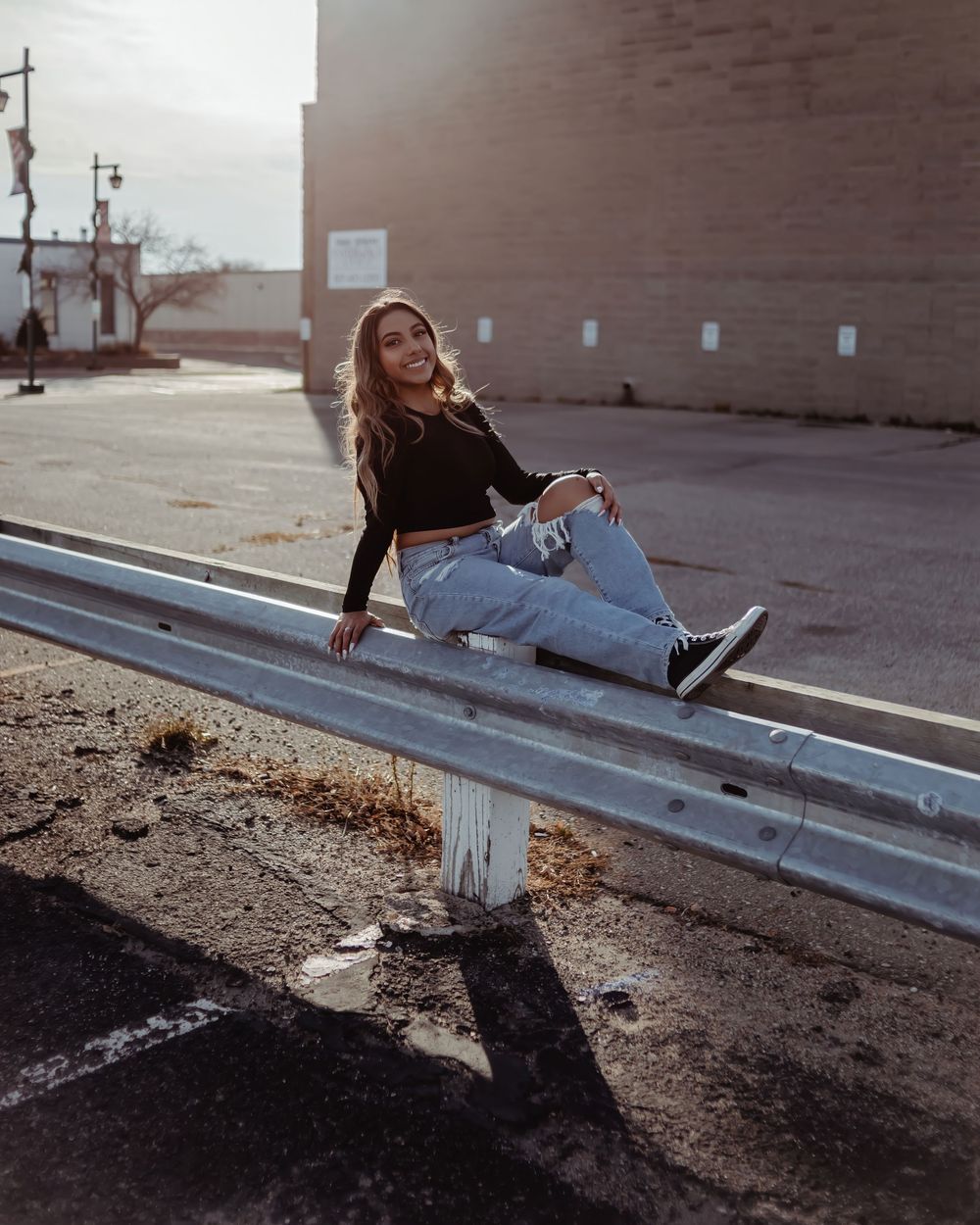 A person in jeans and black top sits casually on a metal guardrail during golden hour lighting.