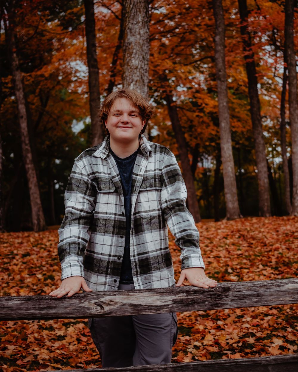 Someone in a plaid flannel jacket leans against a wooden fence surrounded by autumn trees with vibrant orange leaves.
