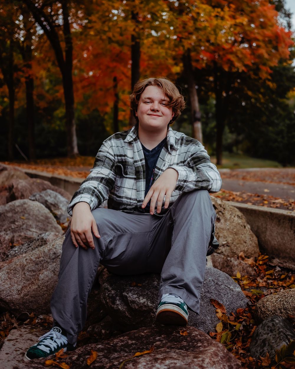Portrait of someone in a plaid shirt sitting on rocks surrounded by vibrant autumn foliage in a forest setting.