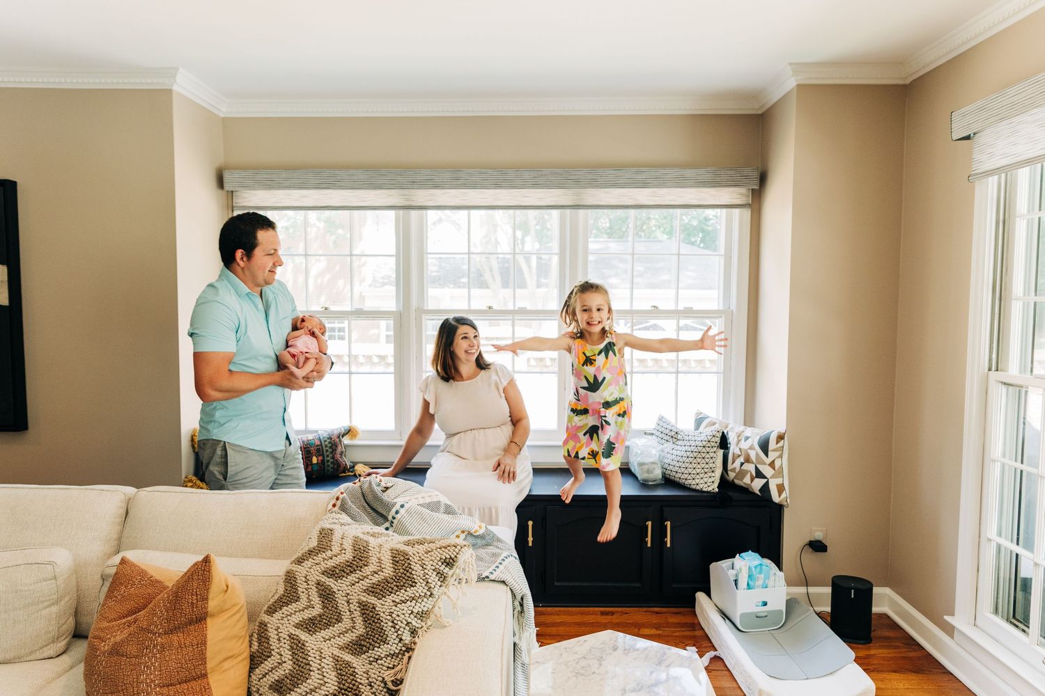 A group gathers together in a sunny living room with large windows, enjoying a light-hearted moment on the couch.
