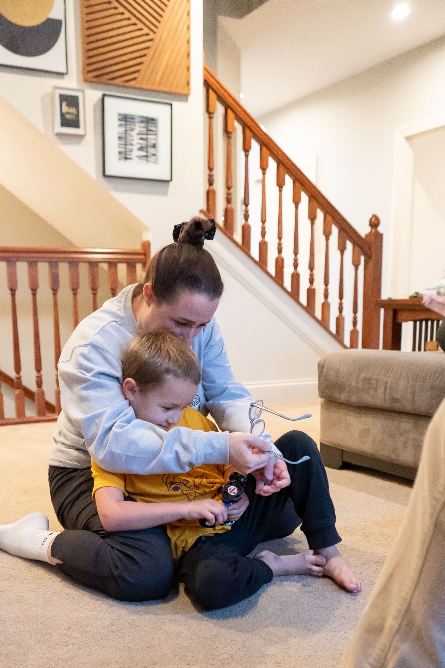 Child sitting on light beige carpet next to wooden staircase plays with a small toy while wearing casual clothing.