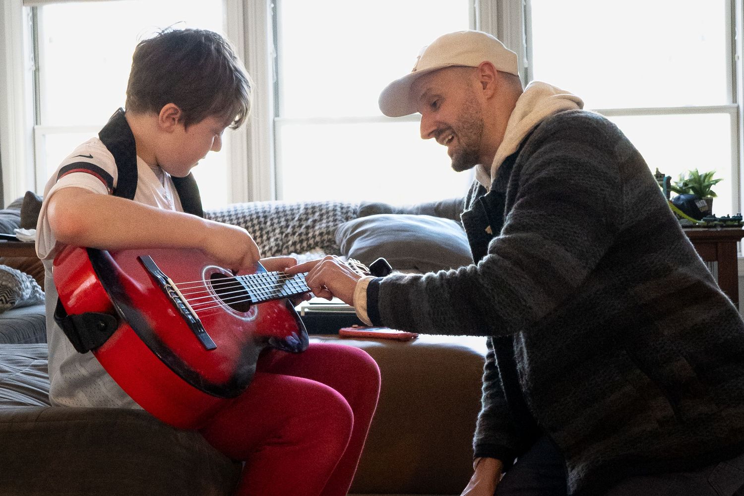 an adult teaching a child how to play guitar while sitting on a couch in a bright room with large windows.