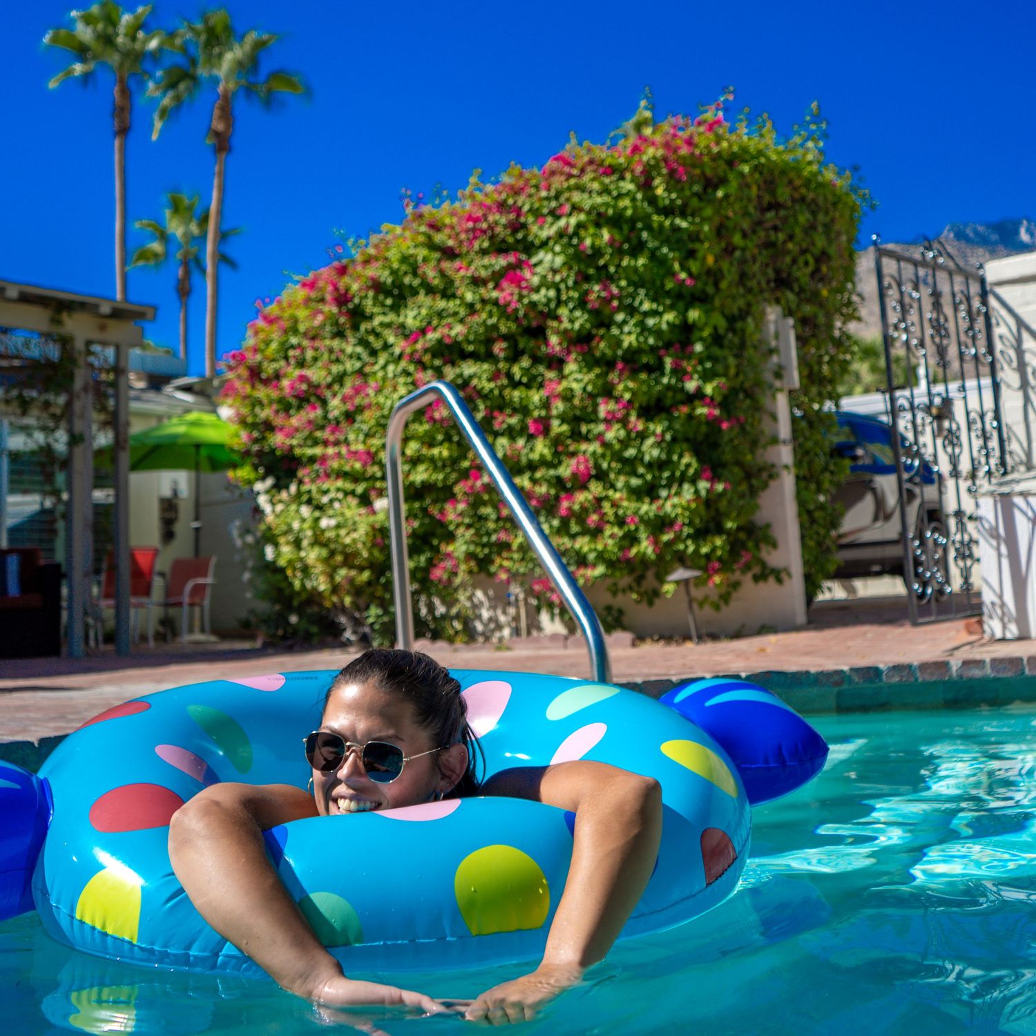 Person floating in blue pool water on colorful inflatable ring with palm trees and flowering shrubs in background.