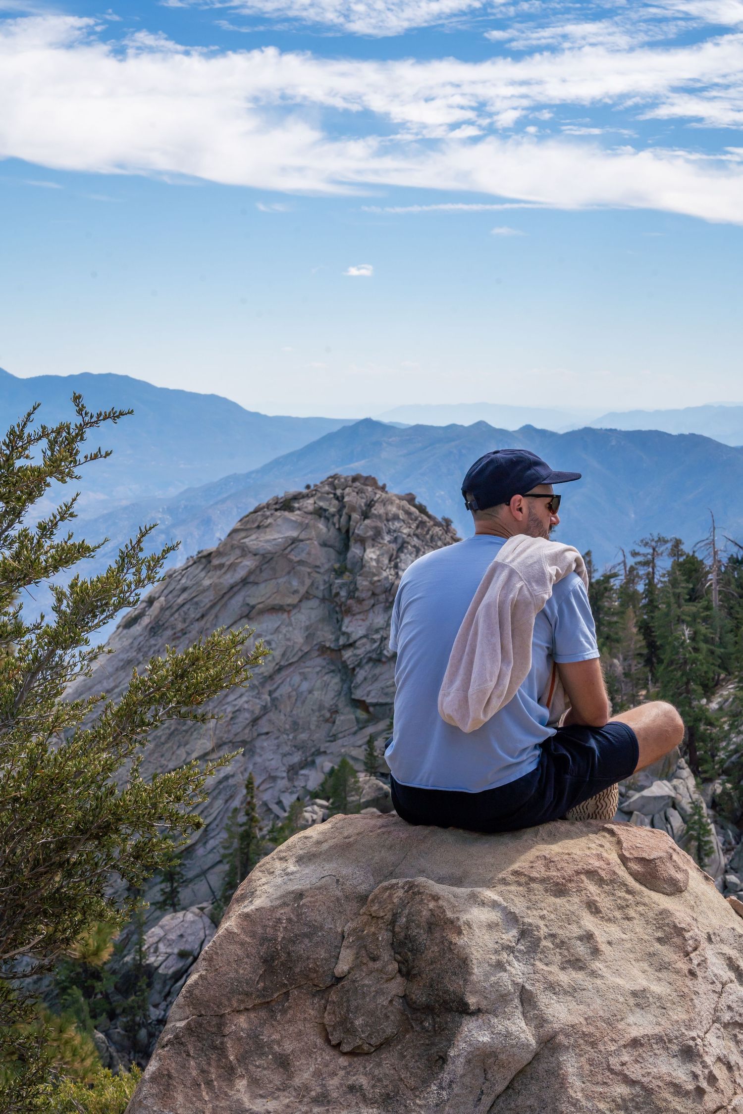 Person in blue shirt sits peacefully on San Jacinto Mountain in California overlooking distant mountain ranges under blue sky.