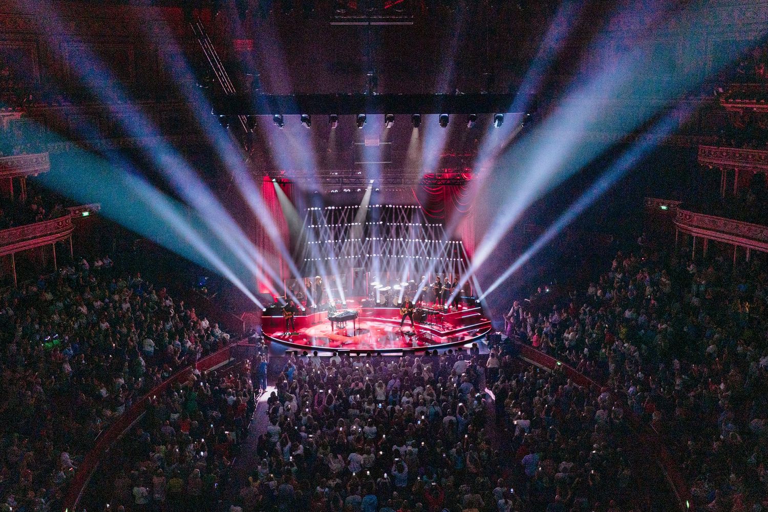 Stadium concert venue bathed in blue and red theatrical lighting with laser beams cutting through atmospheric haze.