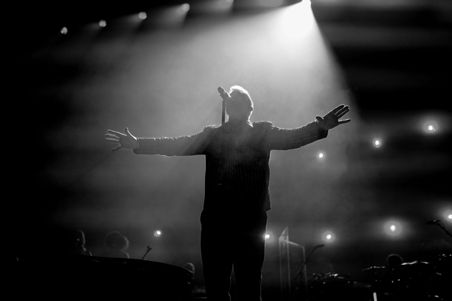 Silhouetted person performs with arms outstretched on stage with dramatic backlighting and fog effects during concert.