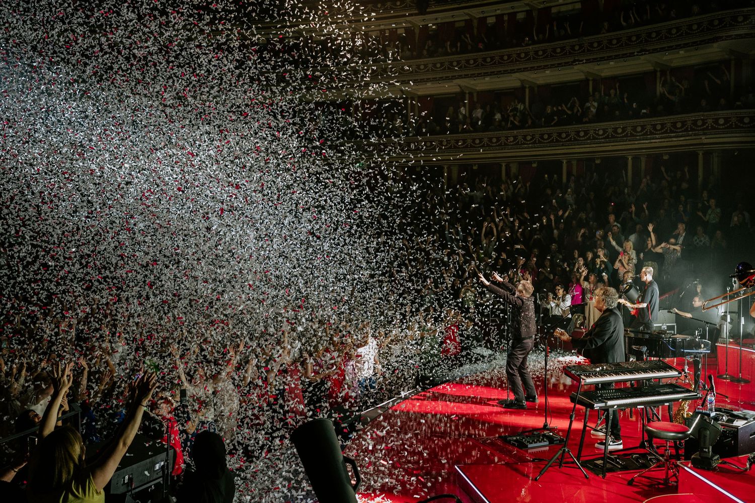 Arena crowd enjoying a concert performance with red and white confetti raining down.
