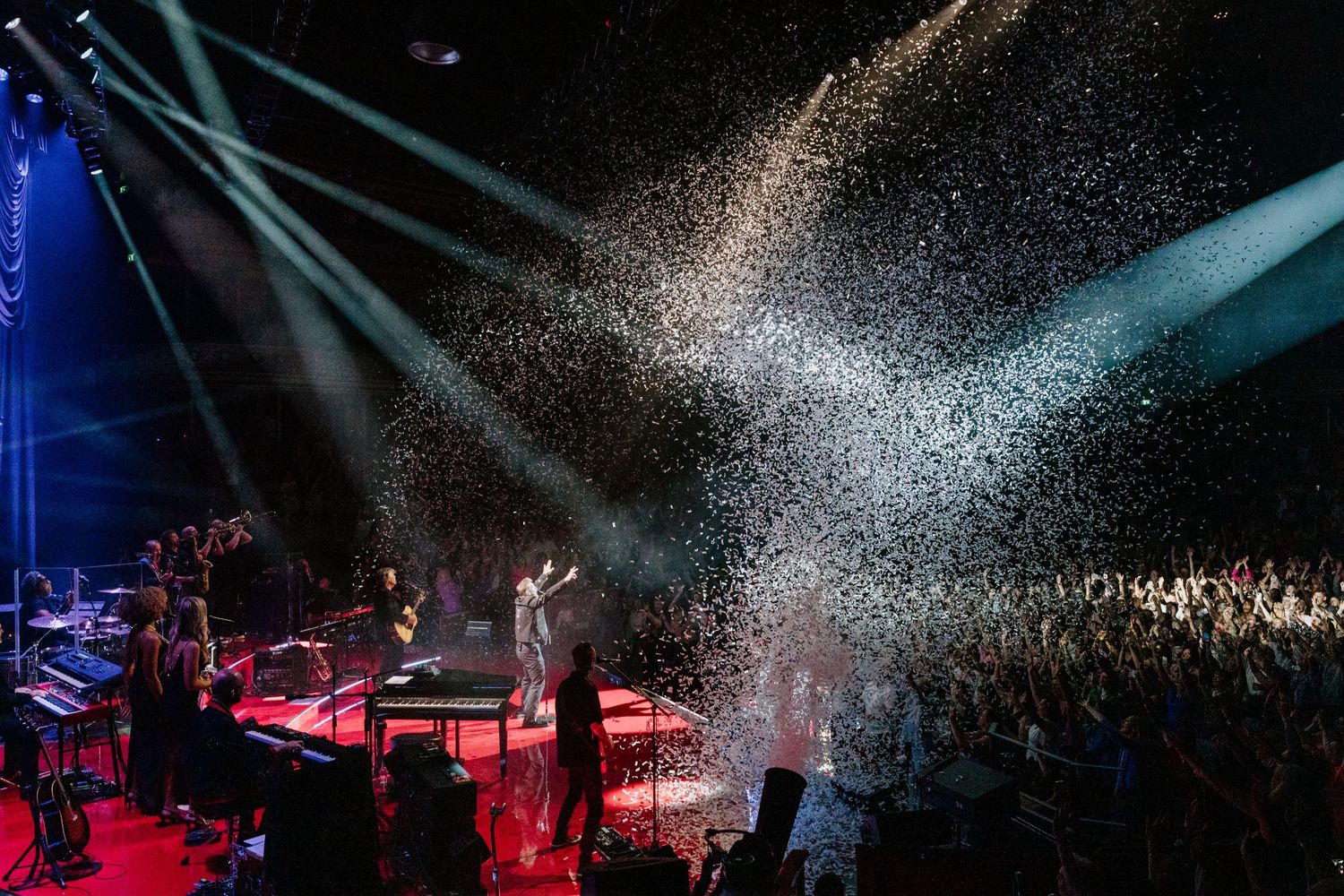Performers on stage during a concert with dramatic red and blue lighting effects creating an atmospheric ambiance.