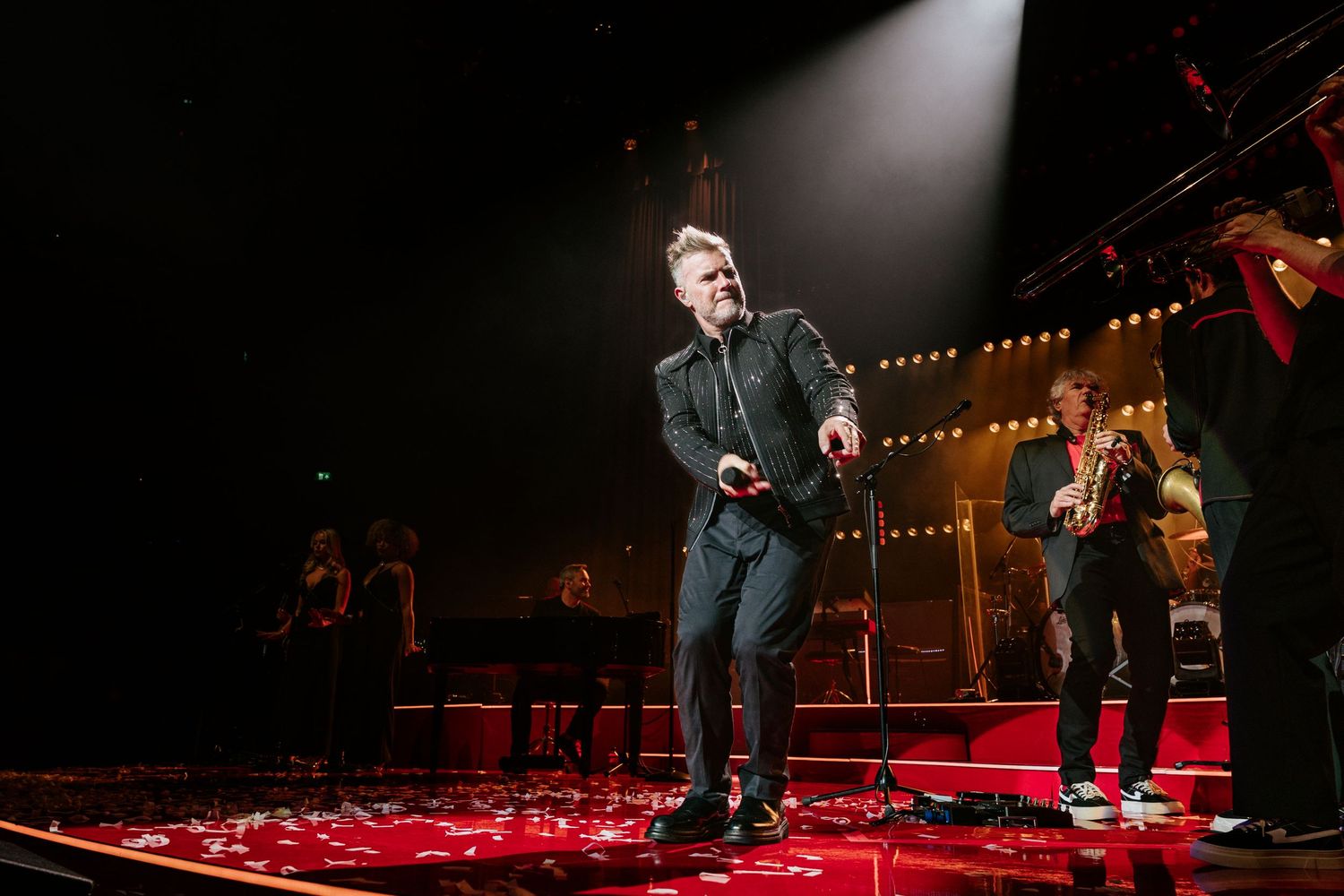 A performer sings on a brightly lit concert stage with red carpet flooring and spotlights illuminating the scene.