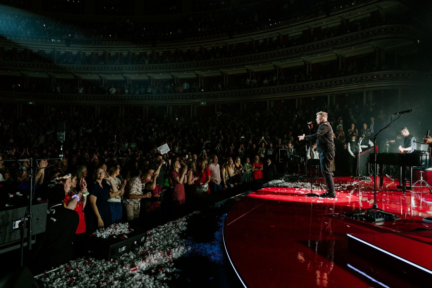 Dark concert stadium illuminated by red spotlights showing multiple performers on a large stage during a performance.