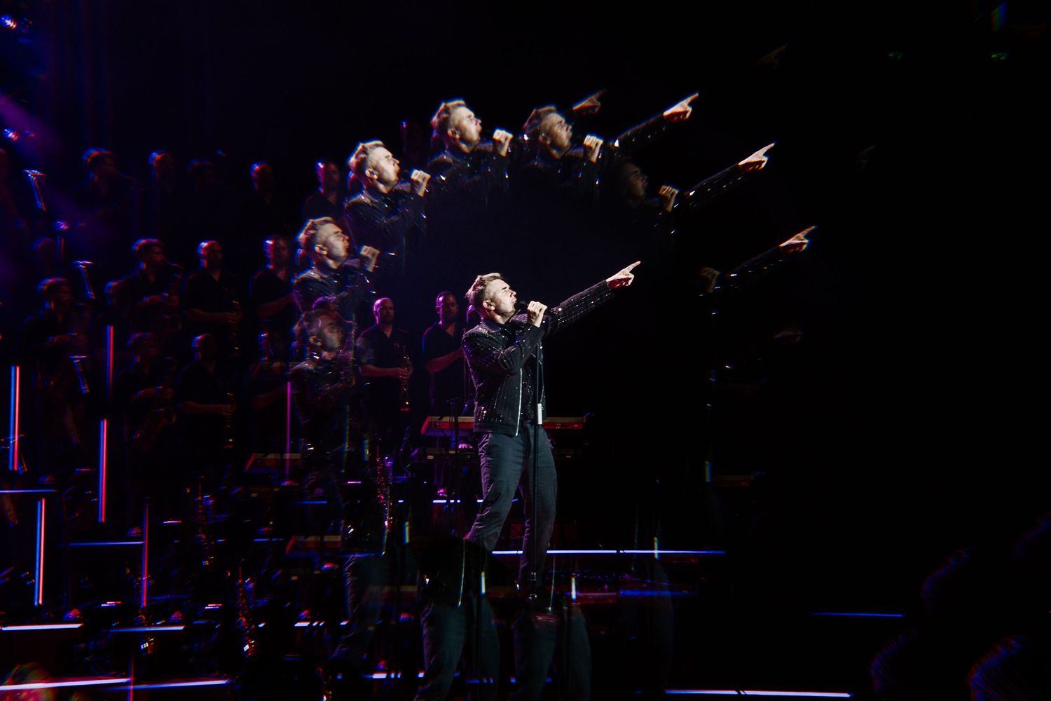 A performer in a dark stage setting gestures during a concert performance with dramatic lighting effects.