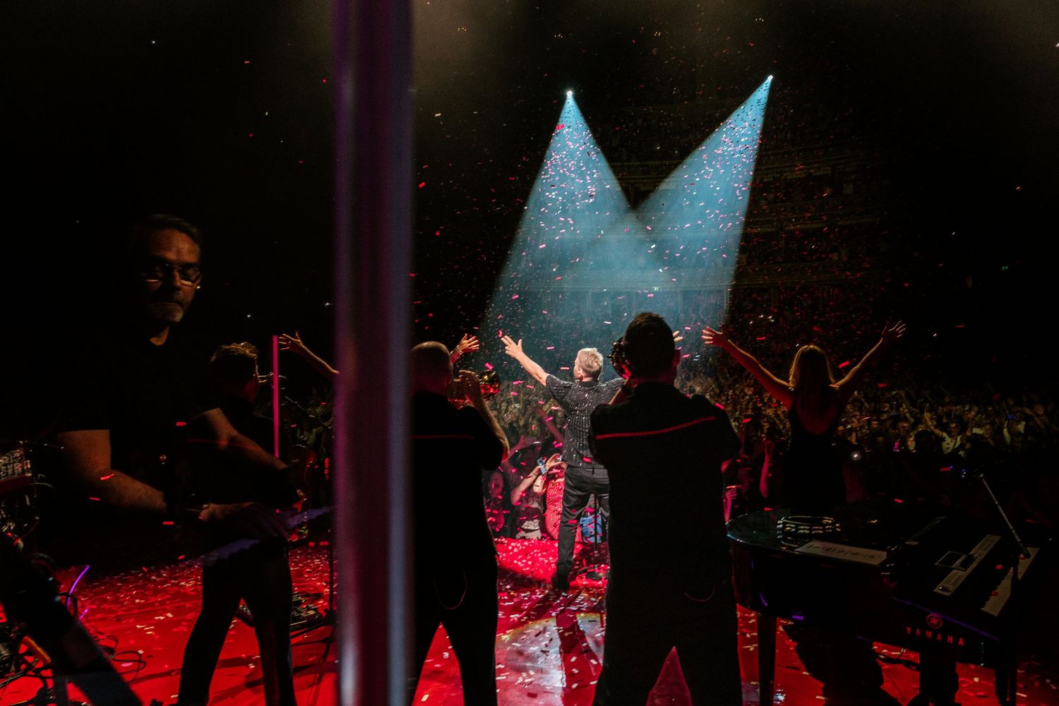 Silhouettes of performers on a dimly lit stage with dramatic red lighting effects in a large venue.