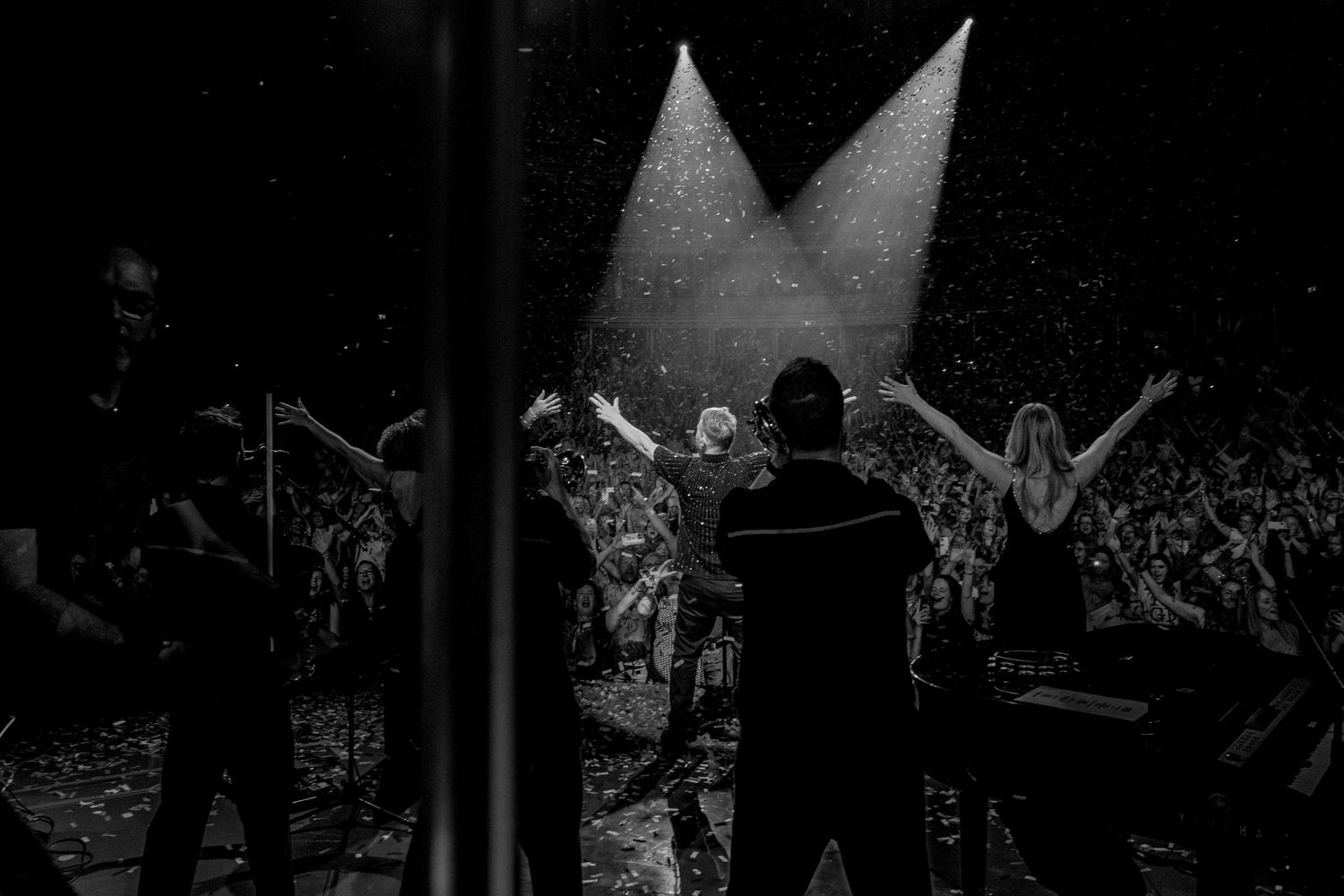 Black and white concert photo showing silhouetted performers on stage with dramatic spotlights and crowd.