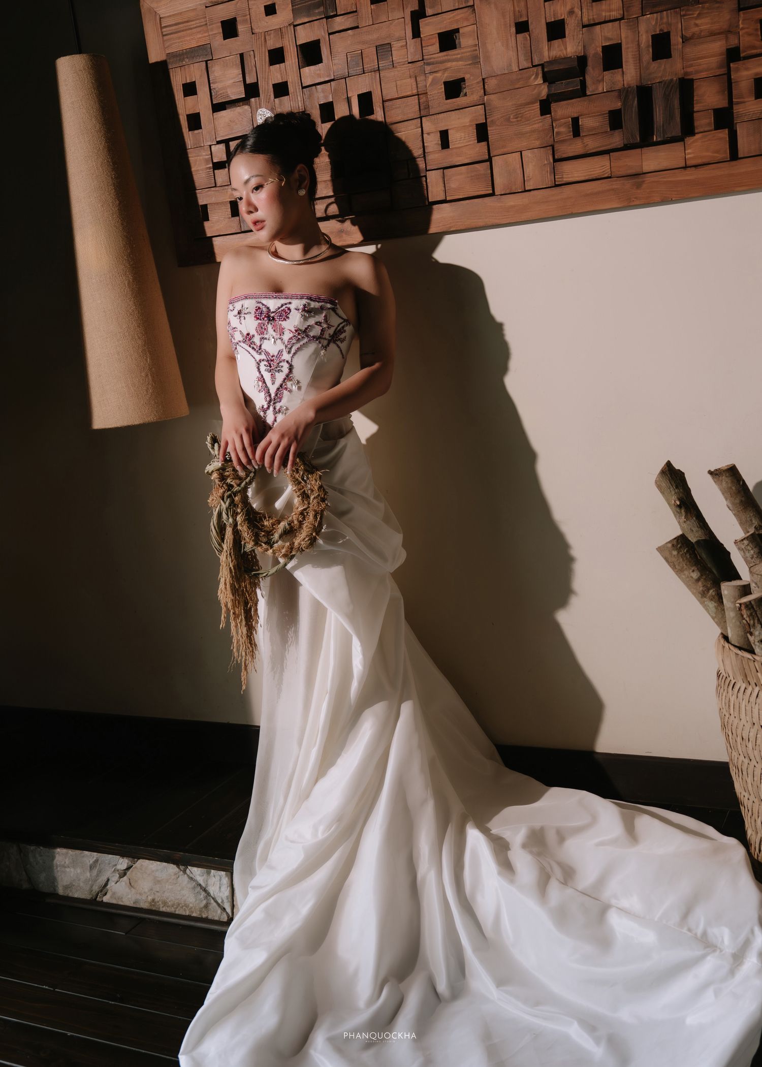 Elegant bride in white embroidered wedding gown holding dried floral wreath in warm light.
