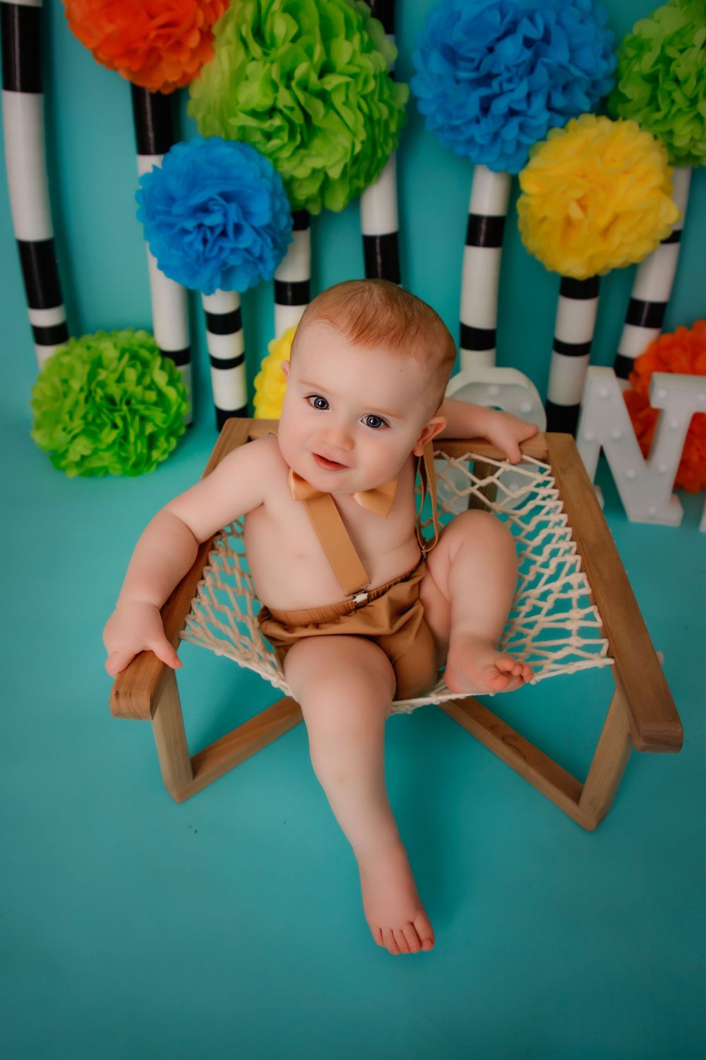 Baby in brown outfit sits in wooden chair against turquoise wall decorated with colorful tissue paper pom poms.