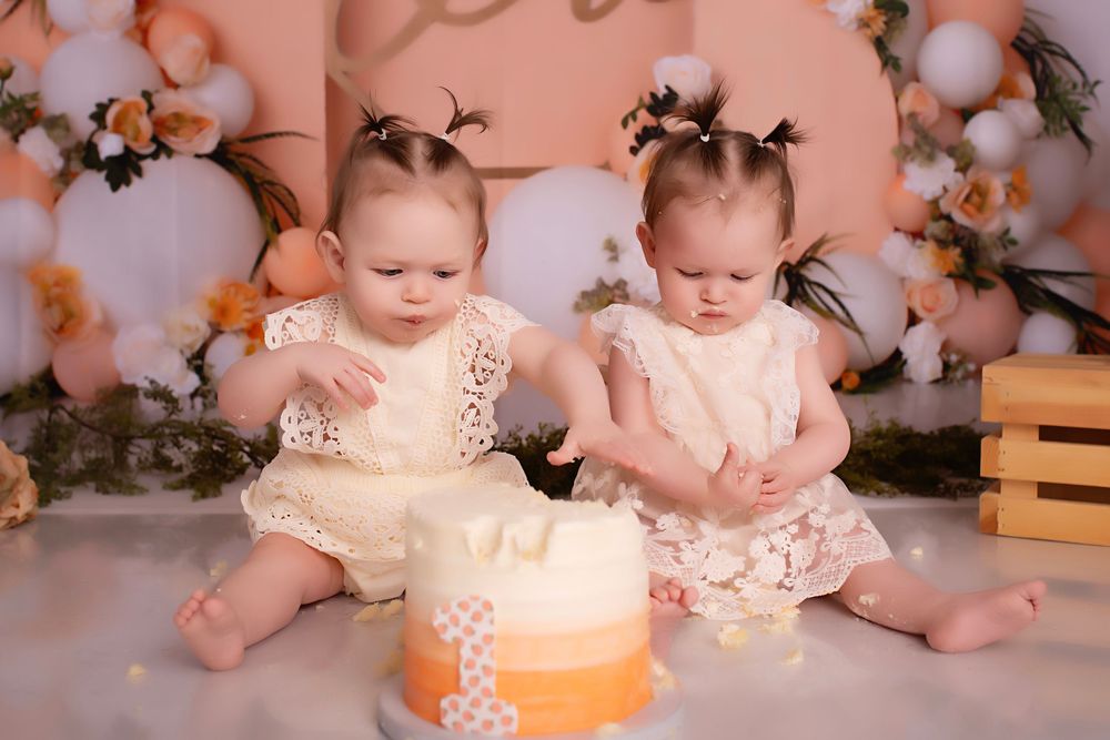 Small children in peachy pink outfits sit together with decorative lights and cotton flowers in background.