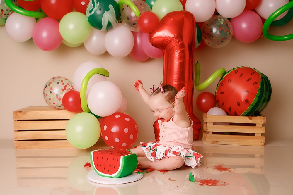 A festive balloon garland with pink, red, green and white balloons featuring a watermelon theme against a tan backdrop.
