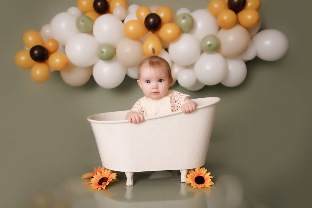 Small baby poses in a vintage white bathtub decorated with orange sunflowers and balloon garland.