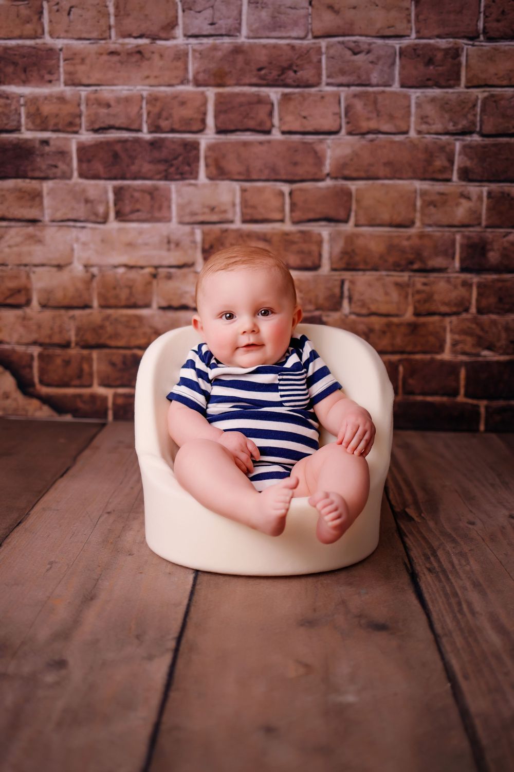 Three photo sequence shows infant wearing navy striped outfit nestled in cozy cream-colored support cushion on wooden floor.
