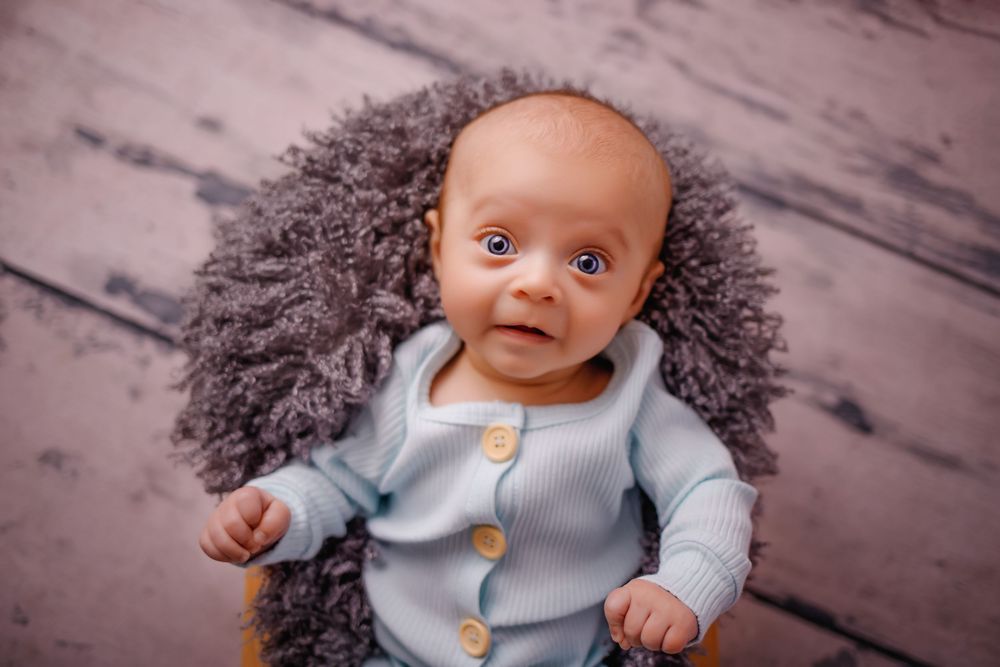 A baby in a light blue sweater sits with curly hair against a rustic wooden backdrop.